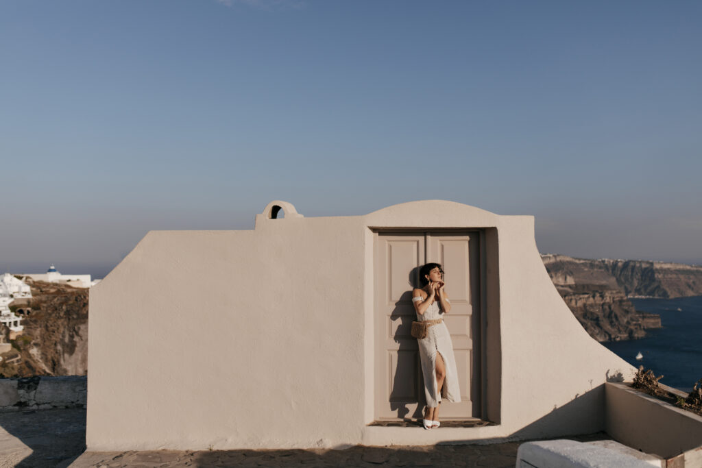 Woman in a white dress standing beside a whitewashed Santorini building overlooking the Aegean Sea