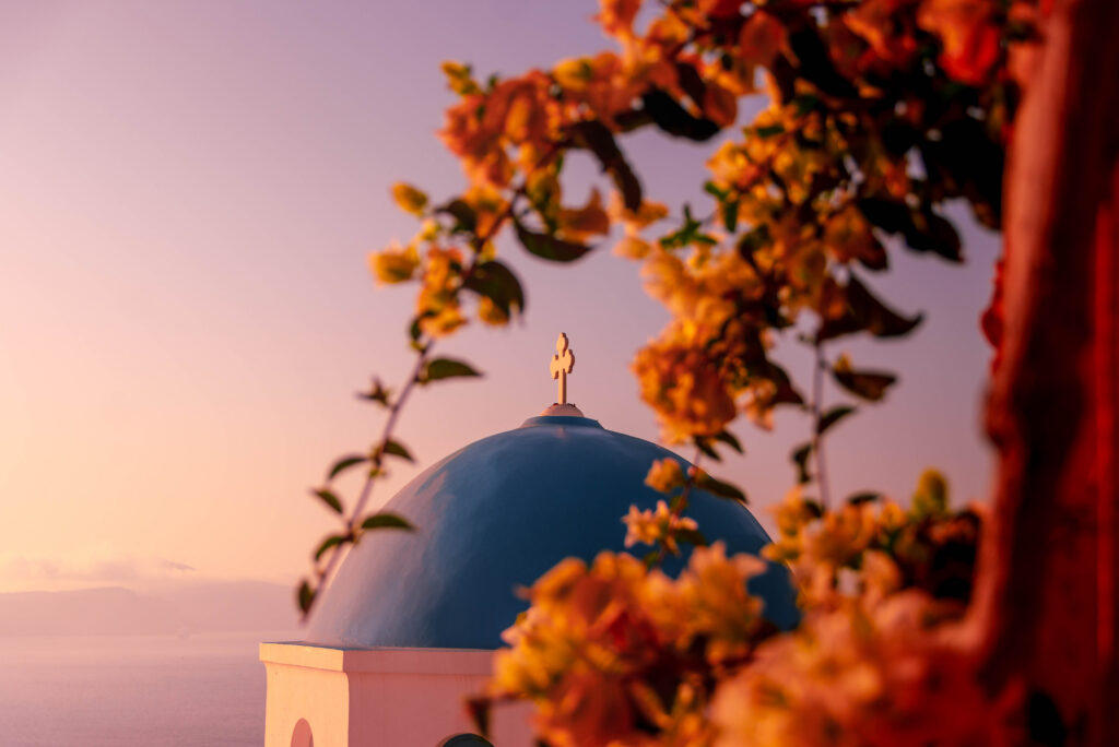Blue-domed church at sunset framed by orange bougainvillea flowers overlooking the Aegean Sea in Greece